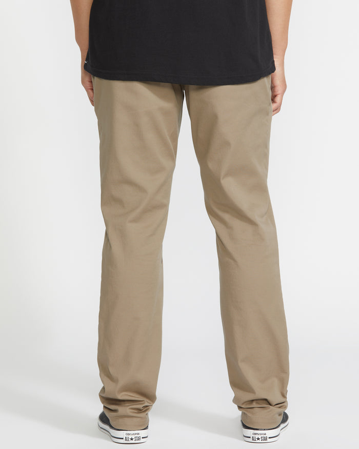 A person wearing Volcom Men’s Frickin Modern Stretch Pants, a black shirt, and white sneakers stands facing away from the camera against a white background.