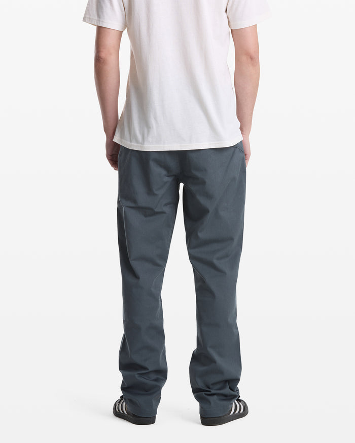 A person stands with their back to the camera, wearing a white t-shirt, Volcom Men's Frickin Modern Stretch Pants, and black sneakers with white stripes, against a plain white background.