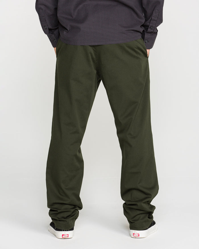 A person stands facing away, wearing Volcom Men's Frickin Modern Stretch Pants in olive green—crafted with REPREVE recycled fibers—paired with a dark long-sleeve shirt and white sneakers featuring red accents, set against a plain light background.