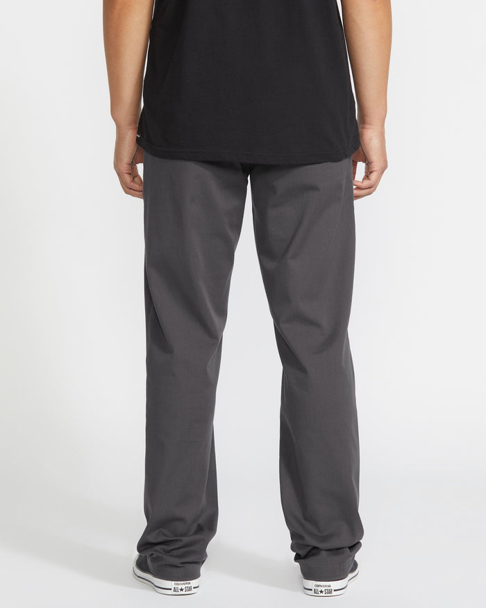 A person stands with their back to the camera, wearing a black t-shirt, Volcom Men's Frickin Modern Stretch Pants made from stretch twill and REPREVE recycled polyester, and white sneakers with black accents, against a white background.