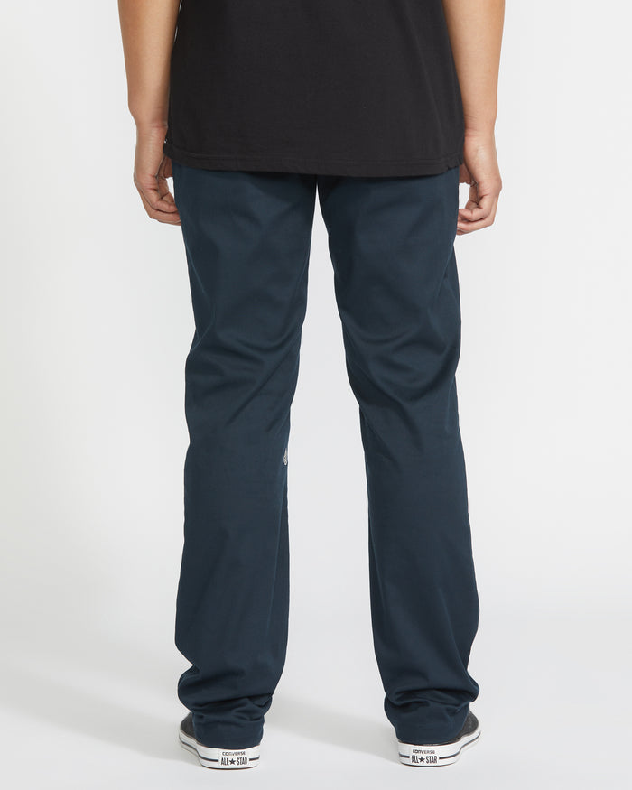 A person stands facing away from the camera, wearing a black shirt, white sneakers, and Volcom Men's Frickin Modern Stretch Pants (slim-fit, five-pocket) against a plain white background.