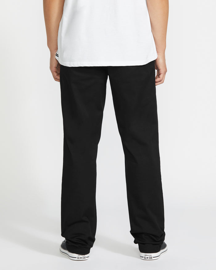 A person faces away from the camera, wearing a white t-shirt, Volcom Men's Frickin Modern Stretch Pants, and black-and-white sneakers against a plain white background.