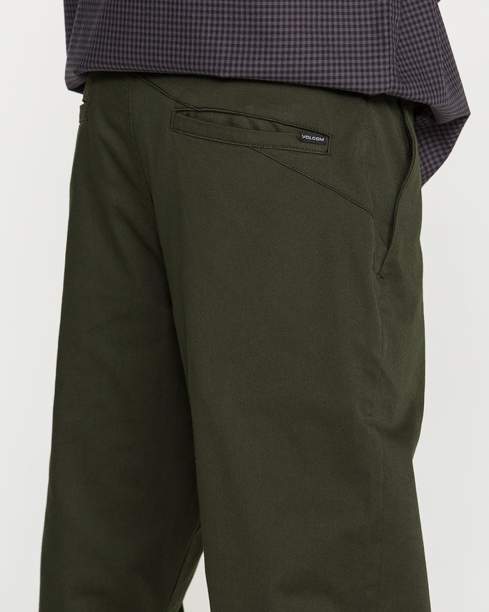 Close-up of a person wearing Volcom Men's Frickin Modern Stretch Pants in dark green, paired with a dark checkered shirt. The back pocket features a small black Volcom label, reflecting the brand’s dedication to quality. Made with REPREVE recycled fibers, these pants deliver both comfort and sustainability. The individual is standing side-on to the camera.