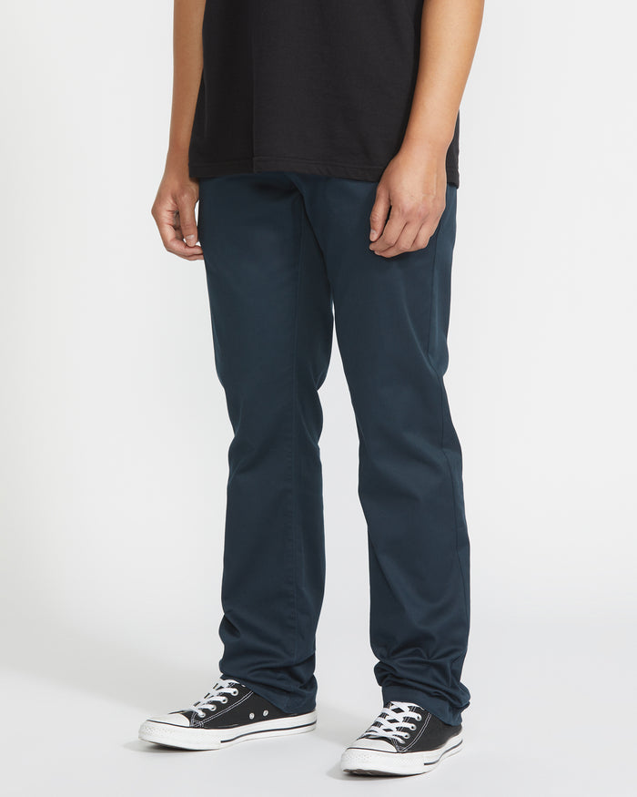 A person stands against a plain white background, cropped from the shoulders down, wearing a black t-shirt, black-and-white sneakers, and Volcom Men's Frickin Modern Stretch Pants featuring slim-fit and five-pocket styling.