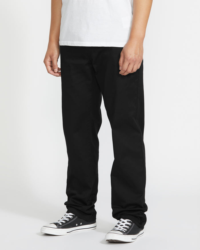 A person, visible only from the waist down, stands against a white background wearing Volcom Men's Frickin Modern Stretch Pants in black, paired with a white T-shirt and black-and-white sneakers.