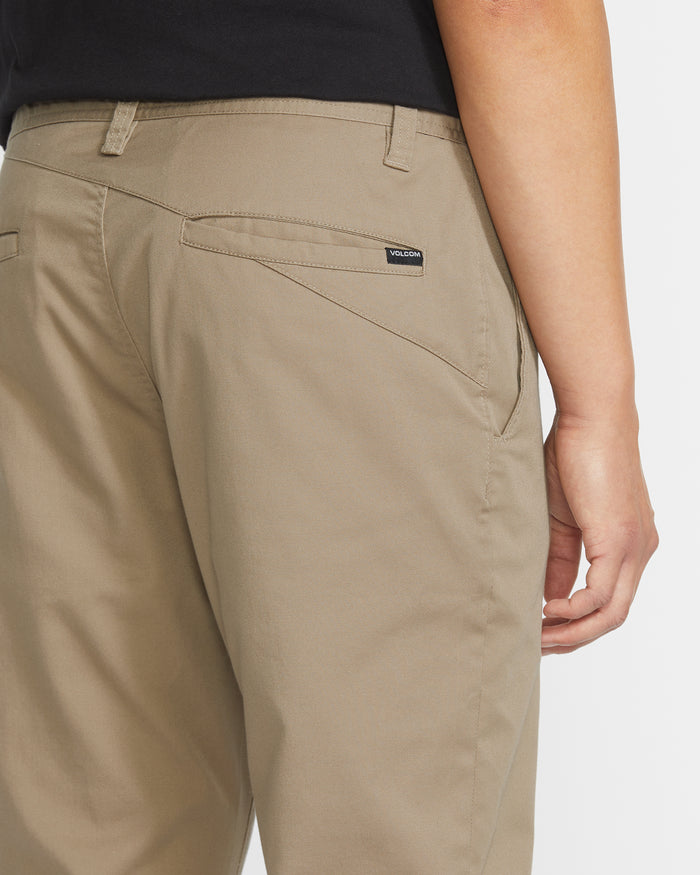 A person is shown from the waist down wearing Volcom Men’s Frickin Modern Stretch Pants in light brown, paired with a black shirt. The pants feature a visible back pocket with a small Volcom brand tag above it and are crafted from recycled polyester for comfort and sustainability.