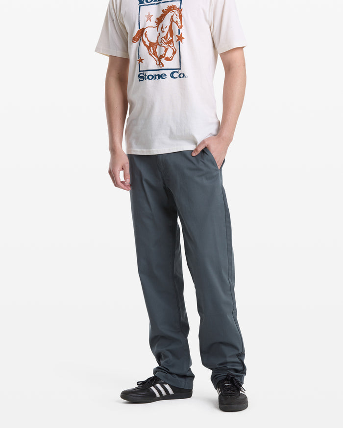 A person wears a white horse graphic tee, Volcom Men's Frickin Modern Stretch Pants in grayish-blue made with REPREVE recycled fibers, and black Adidas sneakers. They stand against a white background with their right hand in their pocket.