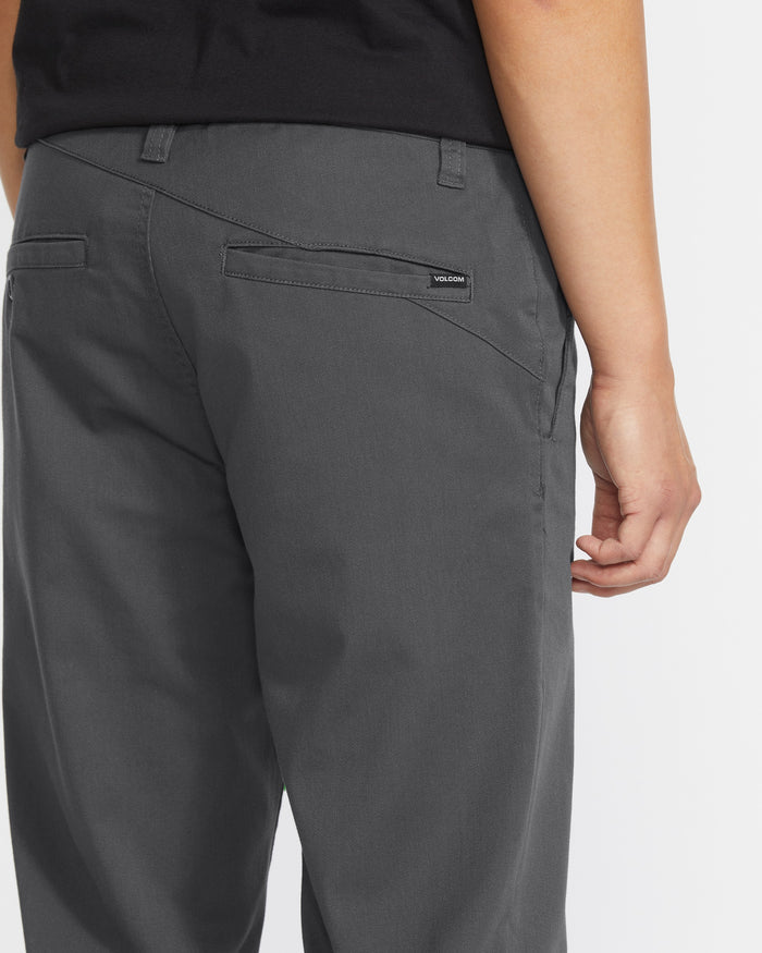 A person is shown from the waist down, facing away, wearing Volcom Men's Frickin Modern Stretch Pants in dark gray and a black shirt. The pants feature a small black Volcom label above the back pocket.