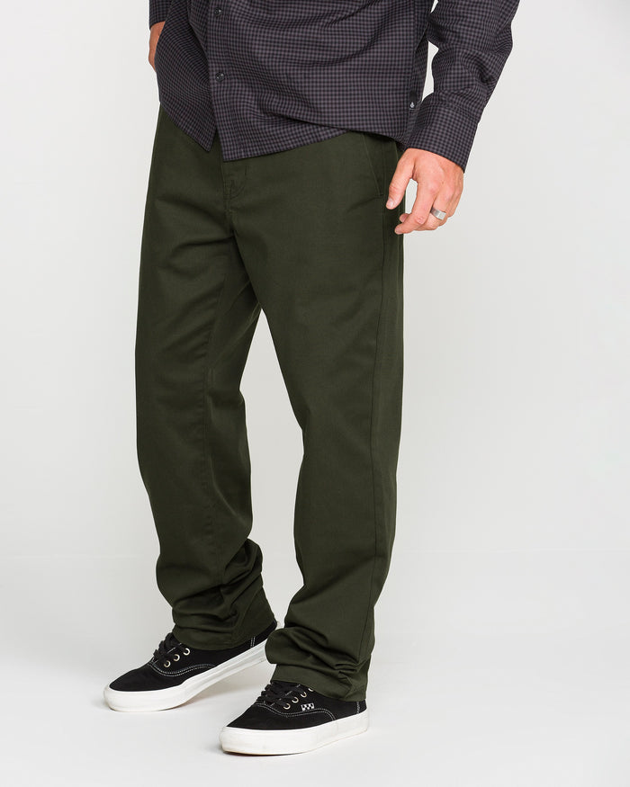 A person wearing a dark checkered shirt, olive green Volcom Men's Frickin Modern Stretch Pants made with REPREVE recycled fibers, and black sneakers with white soles stands against a plain light background.
