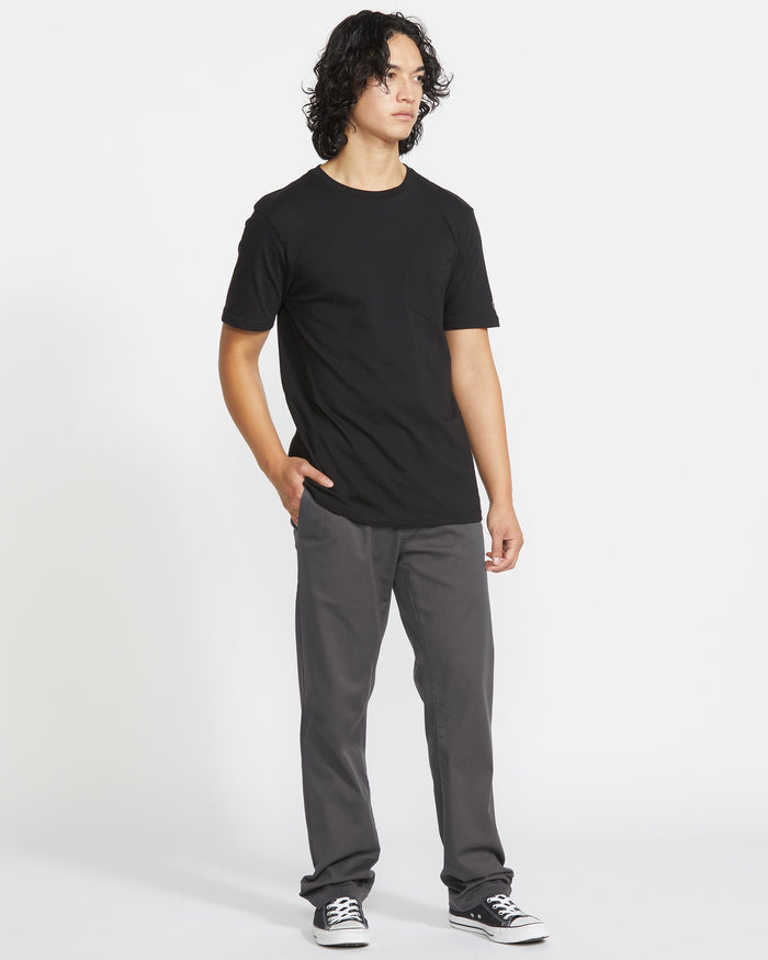 A young man with medium-length curly hair wears a black t-shirt, black sneakers, and Volcom Men's Frickin Modern Stretch Pants in gray. He stands against a plain white background with one hand in his pocket, looking slightly to the side.