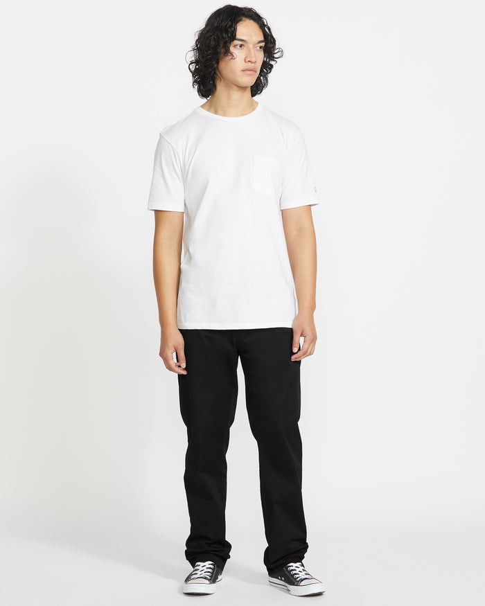 A person with curly hair faces forward against a plain white background, wearing a white T-shirt, black Volcom Men's Frickin Modern Stretch Pants made with REPREVE recycled fibers, and black sneakers with white soles.