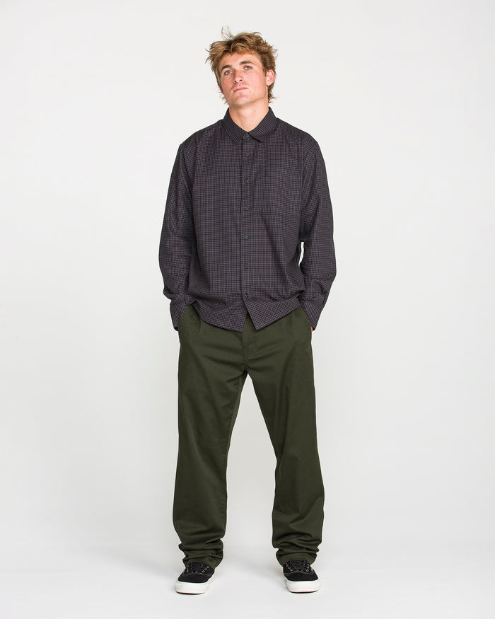 A person stands against a plain white background, wearing a dark checkered Volcom button-up shirt, Volcom Men's Frickin Modern Stretch Pants in olive green made with REPREVE recycled fibers, and black shoes, looking slightly upward with hands in pockets.