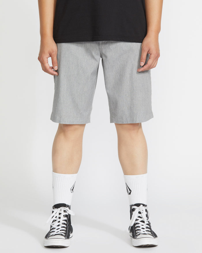 A person wearing a black shirt, Volcom Men’s Frickin Modern Stretch Shorts in light gray, white crew socks with black designs, and black-and-white sneakers stands against a plain white background.
