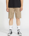 A person stands against a plain white background, wearing a black t-shirt, Volcom Men's Frickin Modern Stretch 21" shorts in khaki, white crew socks with a black logo, and black low-top sneakers.