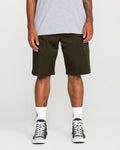 A person stands against a plain white background, shown from the shoulders down, wearing a grey t-shirt, Volcom Men's Frickin Modern Stretch Shorts in dark green—crafted from breathable cotton-poly stretch fabric—paired with white socks and black sneakers.