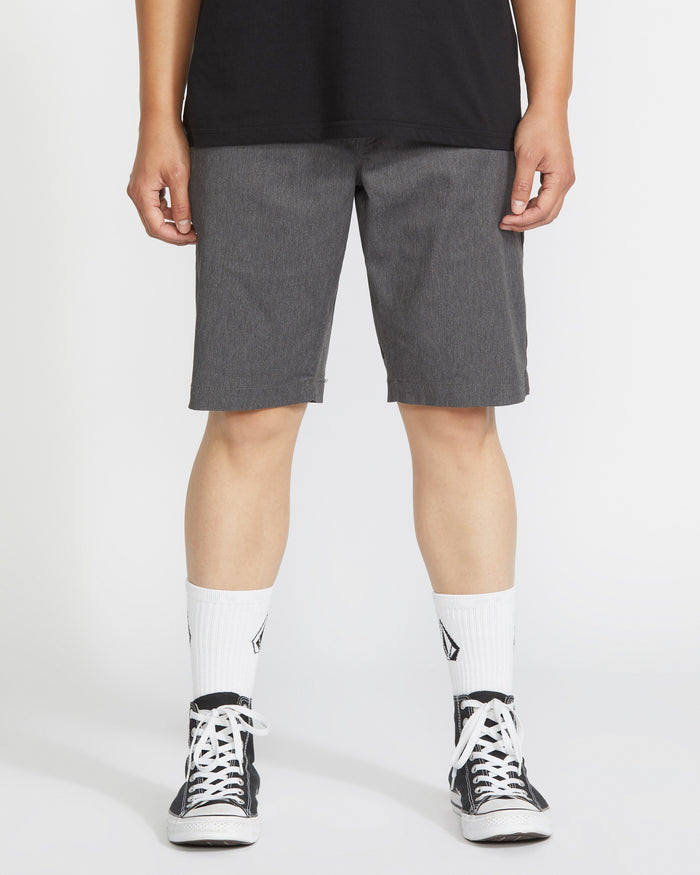 A person wearing a black T-shirt, Volcom Men's Frickin Modern Stretch Shorts in gray, white crew socks with a black logo, and black-and-white sneakers stands against a plain white background.