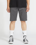 A person wearing a black T-shirt, Volcom Men's Frickin Modern Stretch Shorts in gray, white crew socks with a black logo, and black-and-white sneakers stands against a plain white background.