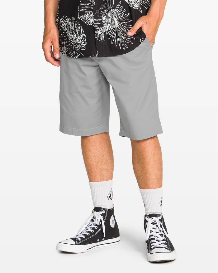 A person wearing Volcom Men's Frickin Modern Stretch Shorts in light gray, a black and white patterned shirt, white socks, and black high-top sneakers stands with one hand in their pocket against a white background.