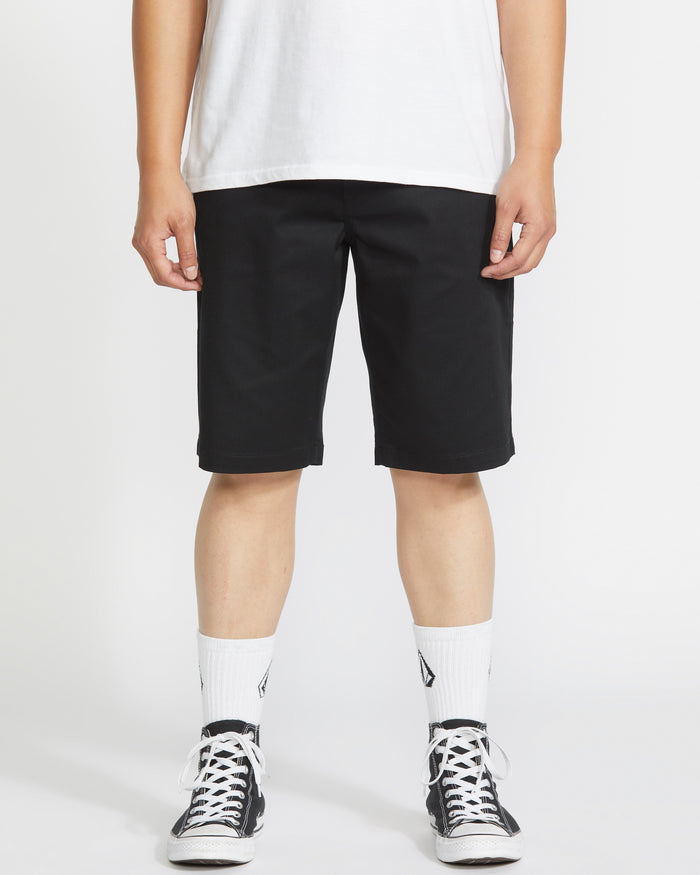 A person stands against a white background, wearing a Volcom white t-shirt, Volcom Men's Frickin Modern Stretch Shorts with 5-pocket styling, white crew socks, and black-and-white sneakers. The image is cropped at the shoulders.