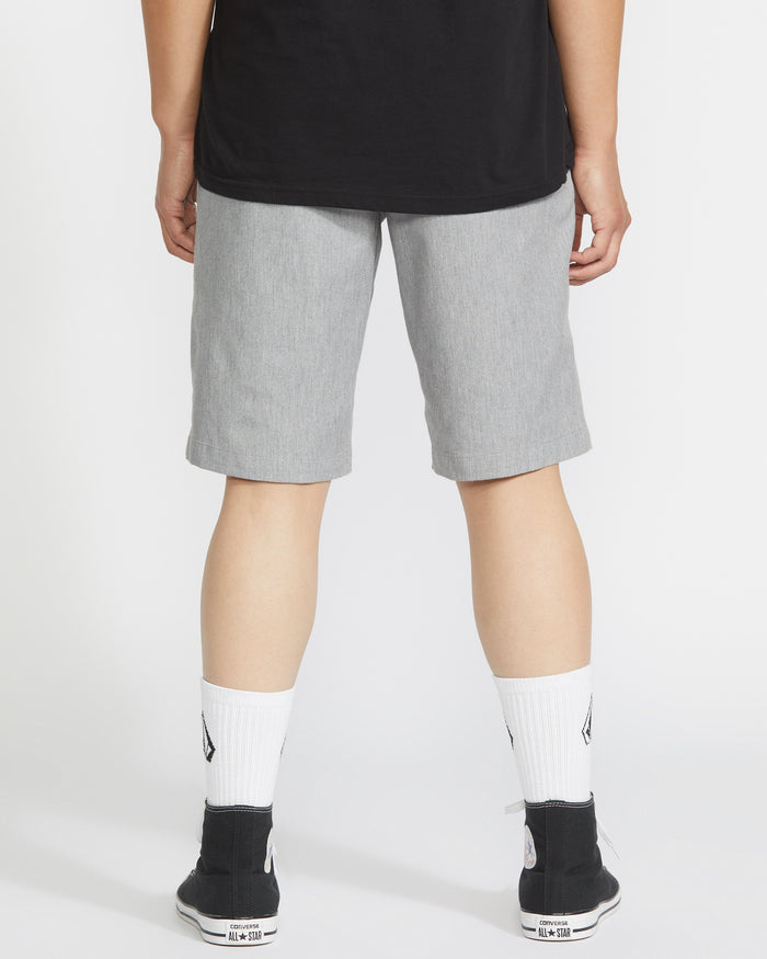A person faces away, wearing a black shirt, Volcom Men's Frickin Modern Stretch Shorts in light gray, white socks with black logos, and black sneakers with white soles, against a plain light background.