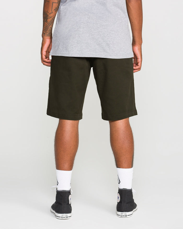 A person stands facing away, wearing a gray t-shirt, Volcom Men's Frickin Modern Stretch Shorts in dark green made from breathable cotton-poly fabric, white socks, and black sneakers.