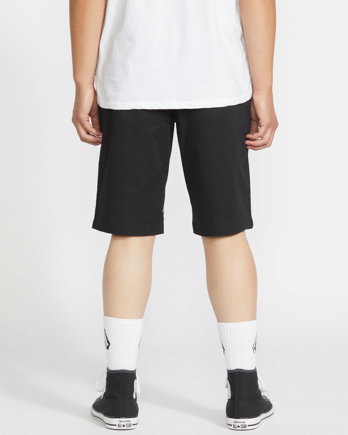 A person is facing away from the camera, wearing a white T-shirt, Volcom Men's Frickin Modern Stretch Shorts with 5-pocket styling, white socks, and black sneakers against a plain white background.