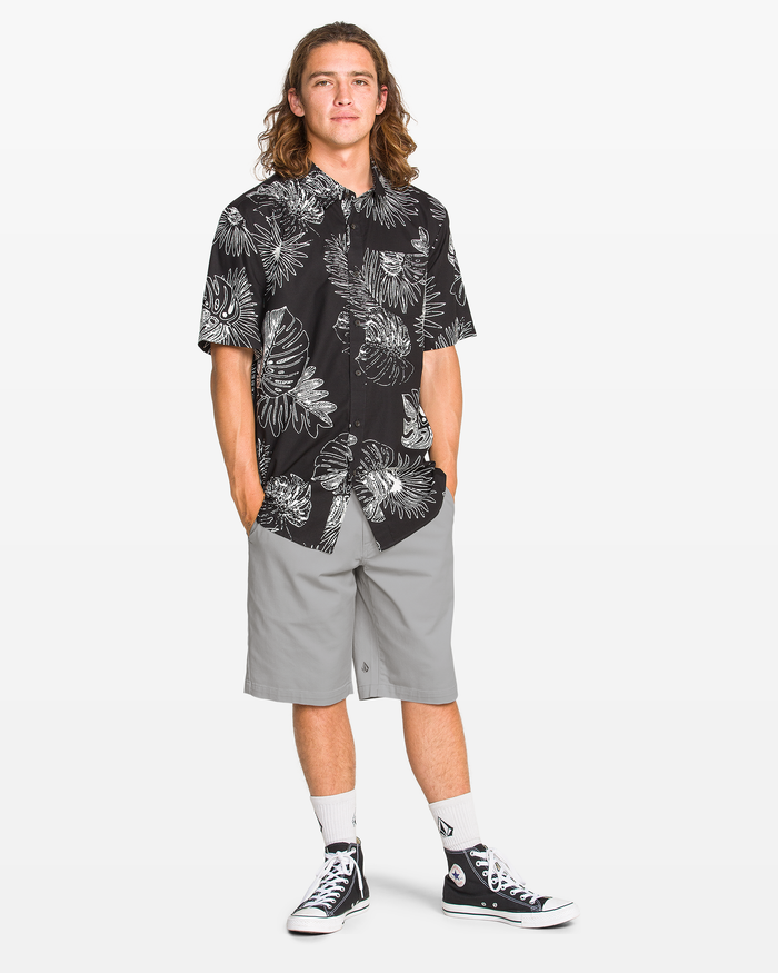 A young man with long hair stands with hands in pockets, wearing a black leaf-patterned shirt and Volcom Men's Frickin Modern Stretch Shorts in light gray, styled with white socks and black Converse sneakers against a plain white background.