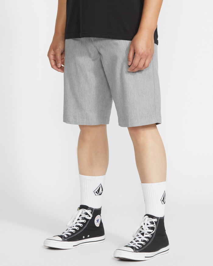 A person in a black t-shirt, white crew socks with a geometric logo, black high-top Converse sneakers, and light gray Volcom Men's Frickin Modern Stretch Shorts stands against a plain white background.
