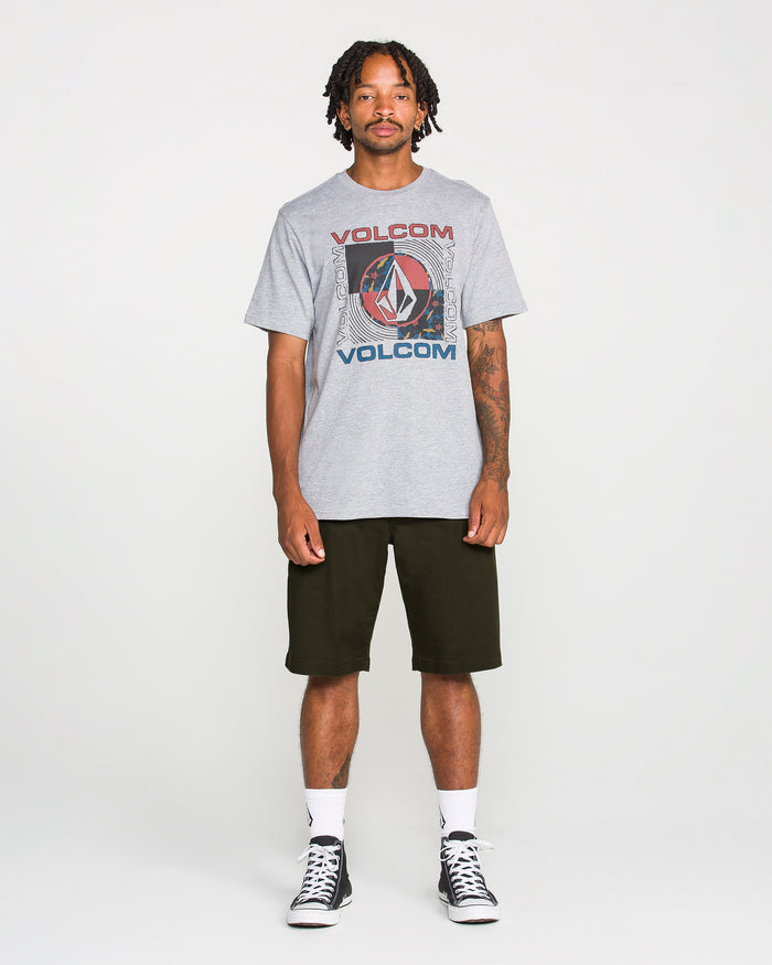 A man stands facing forward against a plain white background, wearing a gray Volcom graphic t-shirt, Volcom Men's Frickin Modern Stretch Shorts in black—crafted from breathable cotton-poly fabric—white socks, and black sneakers.
