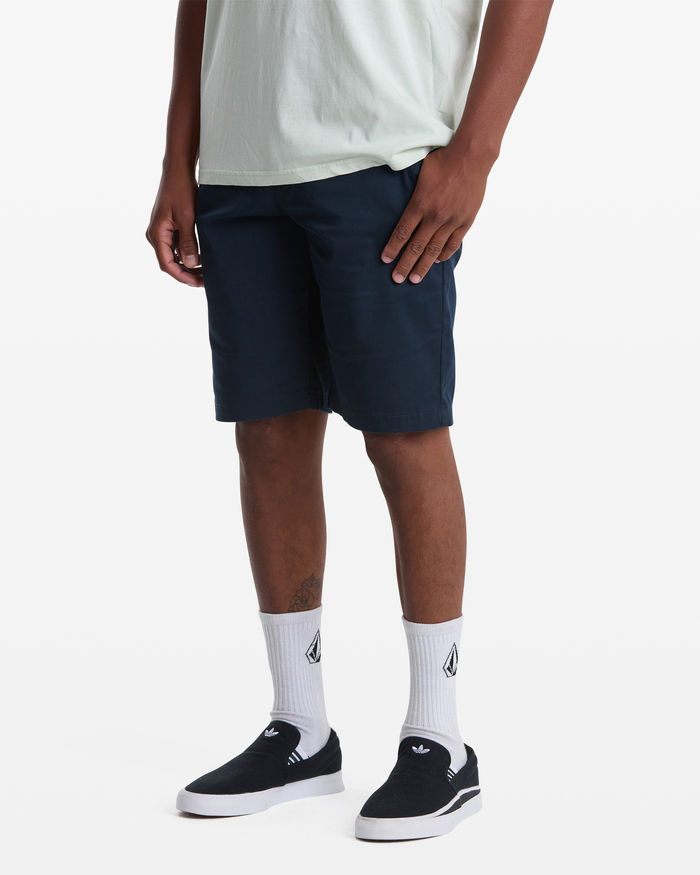 A person with their face not visible stands against a white background, wearing a light-colored t-shirt, Volcom Men's Frickin Modern Stretch Shorts in navy, white socks with black logos, and black slip-on shoes.