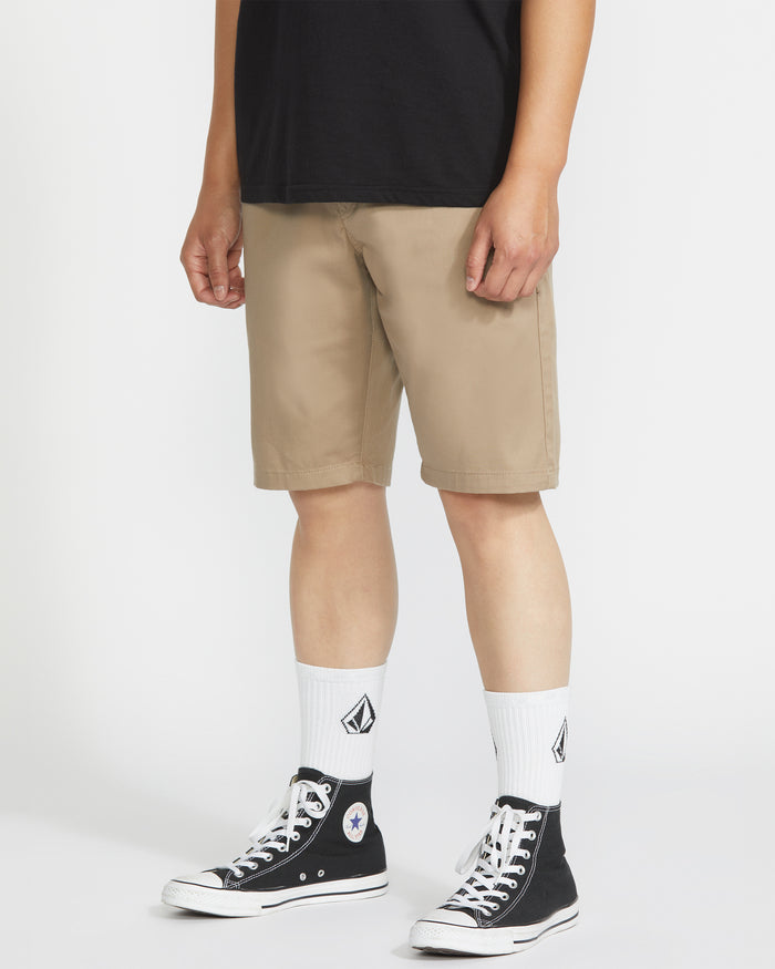 A person wearing Volcom Men's Frickin Modern Stretch 21" Shorts in beige, a black t-shirt, white crew socks with a black logo, and black high-top Converse sneakers stands against a plain white background.