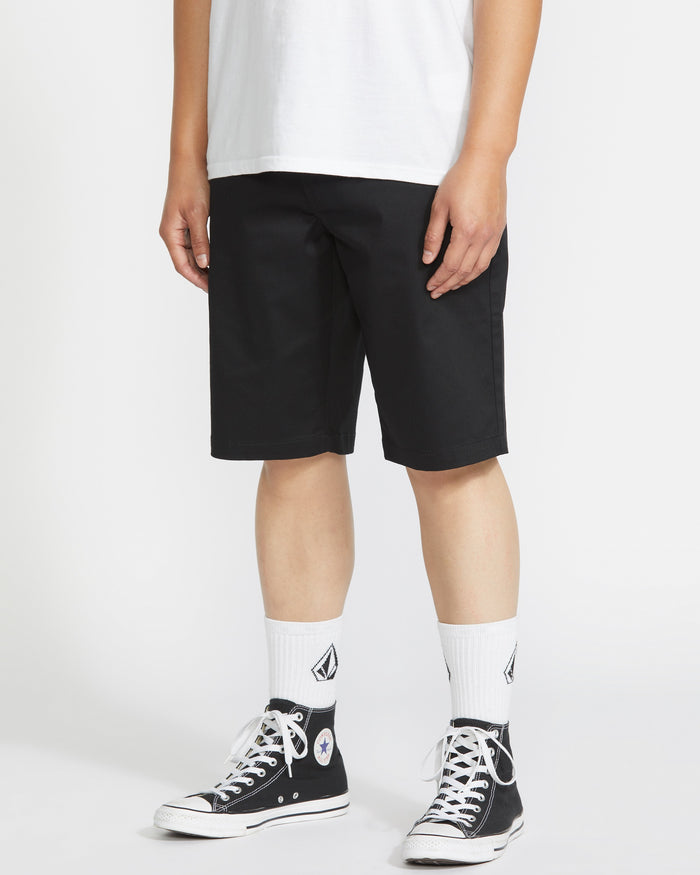 A person wearing a white t-shirt, Volcom Men's Frickin Modern Stretch Shorts, white crew socks with a logo, and black high-top Converse sneakers stands against a plain white background.