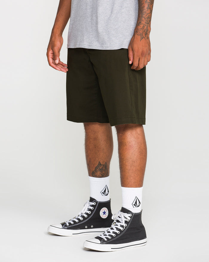 A person wearing a gray t-shirt, Volcom Men's Frickin Modern Stretch Shorts in dark green crafted from breathable cotton-poly fabric, white socks with a black logo, and black high-top Converse sneakers stands against a plain background.