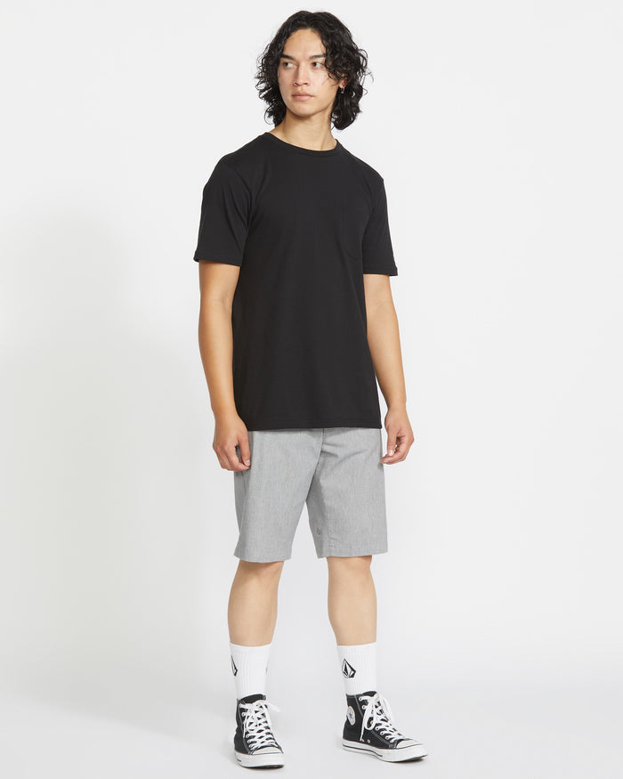 A person with medium-length curly hair wears a black t-shirt, light gray Volcom Men's Frickin Modern Stretch Shorts, white socks with black logos, and black high-top sneakers, standing against a plain white background.