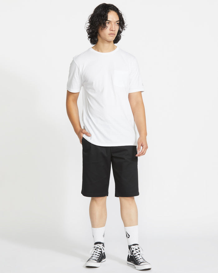 A person with curly hair wears a white T-shirt, Volcom Men’s Frickin Modern Stretch Shorts in black with 5-pocket styling, white socks, and black high-top sneakers, standing against a plain white background.