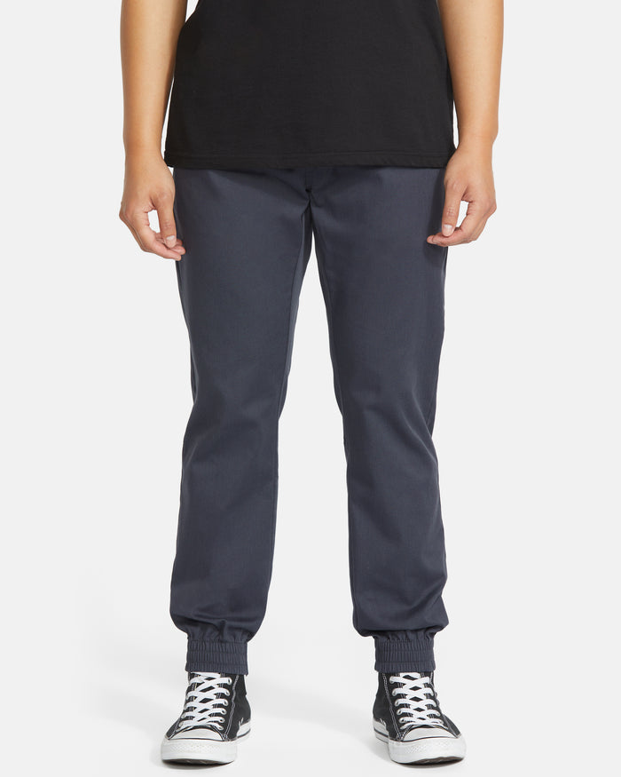 A person wearing a black t-shirt, Volcom Men's Frickin Slim Joggers in dark blue with elastic cuffs, and black-and-white sneakers stands against a plain white background.