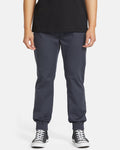 A person wearing a black t-shirt, Volcom Men's Frickin Slim Joggers in dark blue with elastic cuffs, and black-and-white sneakers stands against a plain white background.