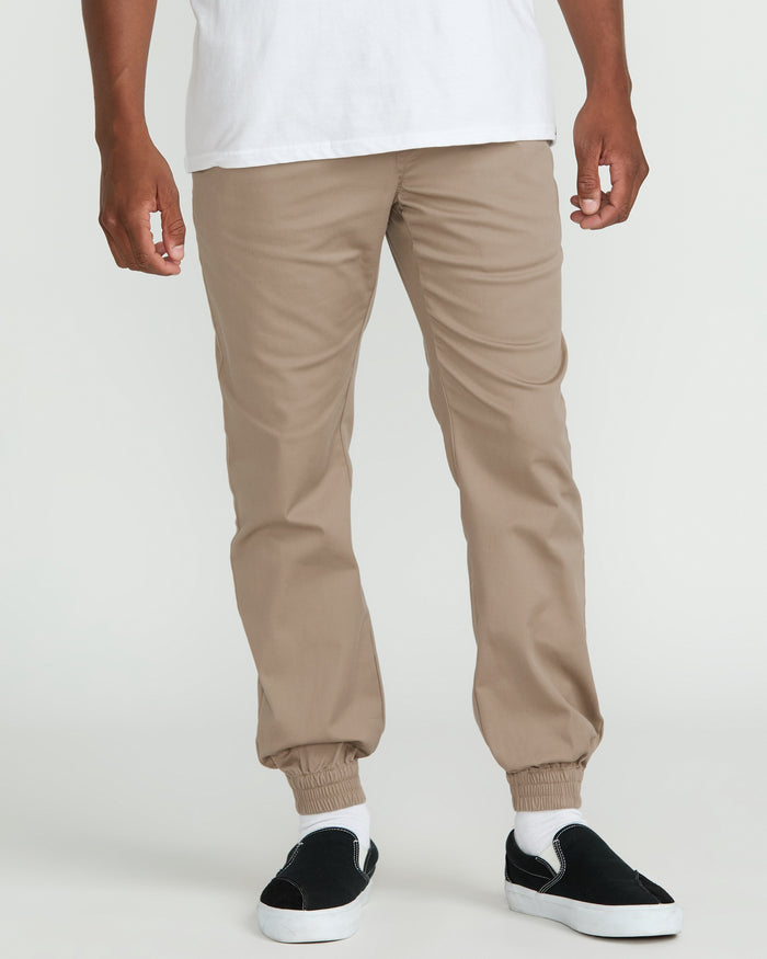 A person wearing Volcom’s Men's Frickin Slim Joggers, made from recycled fibers, paired with a white t-shirt and black slip-on shoes, stands against a plain white background.