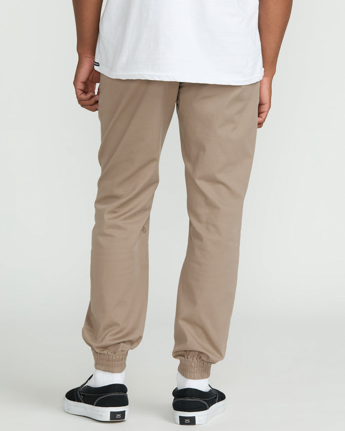 A person stands facing away, wearing a white T-shirt, Volcom Men's Frickin Slim Joggers in tan—crafted from recycled fibers—and black slip-on sneakers with white soles, set against a plain light background.