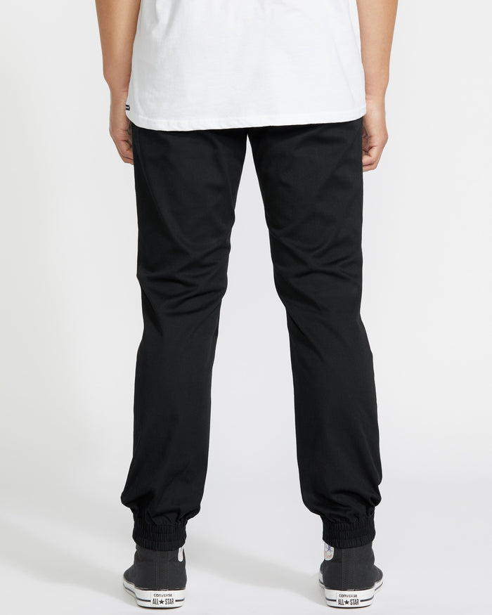 A person stands facing away, wearing a white shirt and Volcom Men's Frickin Slim Joggers featuring elastic cuffs and REPREVE recycled fibers, paired with black and white sneakers. The scene is plain and well-lit.
