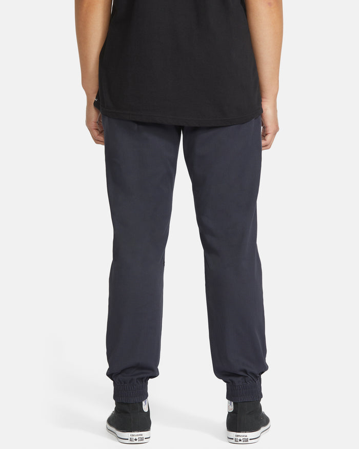 The lower half of a person faces away from the camera, wearing Volcom Men's Frickin Slim Joggers crafted with REPREVE recycled fibers, paired with a black t-shirt and black sneakers with white soles, set against a plain white background.
