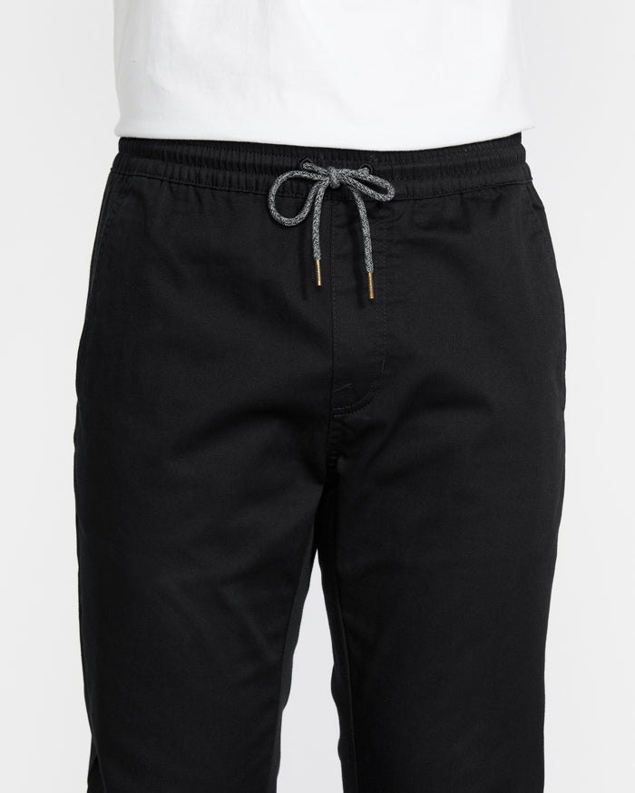 A person is shown from the lower torso down, wearing Volcom Men's Frickin Slim Joggers in black with a visible gray and white drawstring at the waistband, paired with a white shirt. These joggers offer both style and comfort.