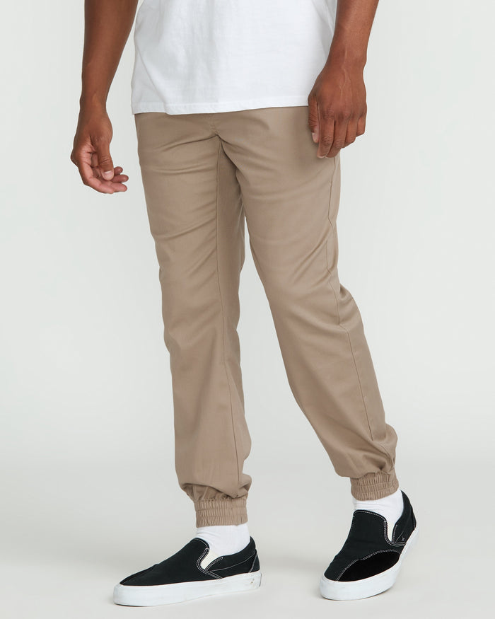 A person wears Volcom Men's Frickin Slim Joggers in tan along with a white T-shirt. Their hands are relaxed at their sides, and they have on black slip-on sneakers with white soles. The background is plain and light-colored.
