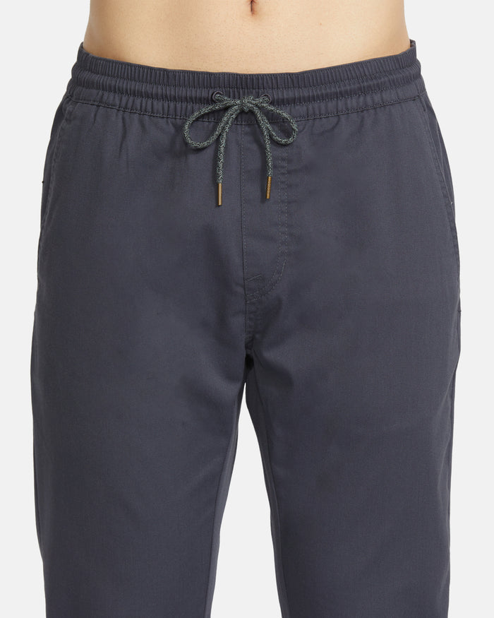 A close-up shows a person wearing Volcom Men's Frickin Slim Joggers in dark gray, featuring an elastic waistband with front drawstring and made with REPREVE recycled fibers.