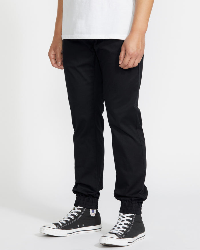 A person stands against a plain white background, cropped from shoulder to feet, wearing a white T-shirt and Volcom Men's Frickin Slim Joggers in black, made with REPREVE recycled fibers, plus black high-top Converse sneakers.