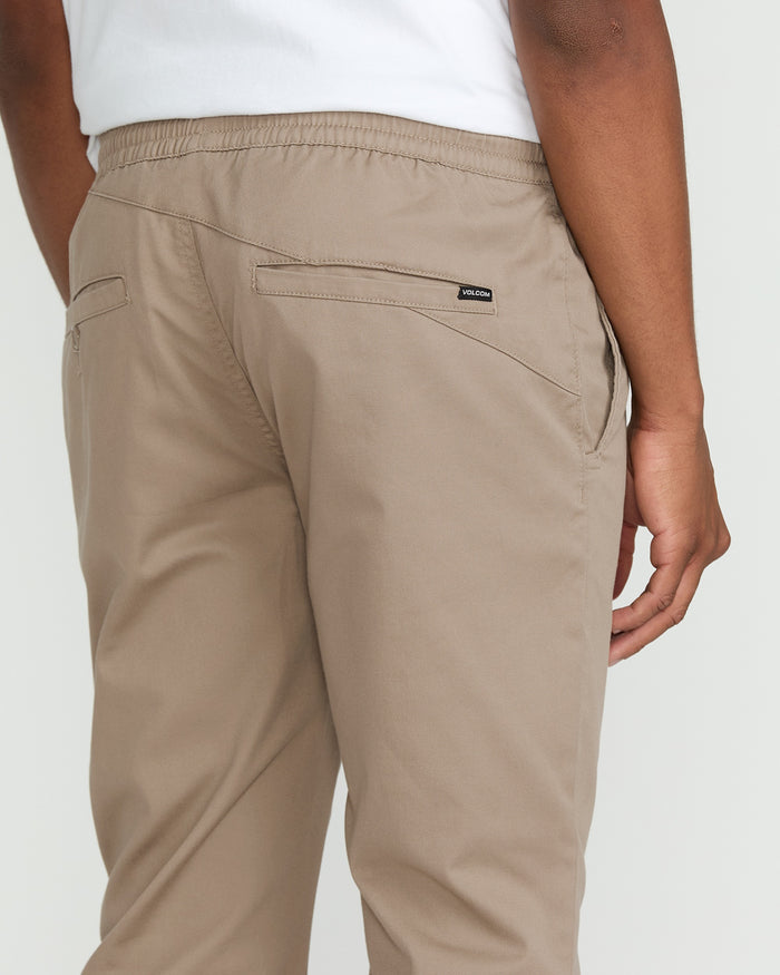 A person is shown from the back wearing Volcom Men's Frickin Slim Joggers in light brown paired with a white shirt. These slim jogger pants feature an elastic waistband, two back pockets, a small black Volcom tag near one pocket, and are thoughtfully crafted from recycled fibers.