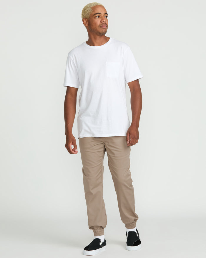 A person with short blond hair wears a white pocket t-shirt, Volcom Men's Frickin Slim Joggers in beige made from recycled fibers, and black slip-on shoes, standing against a plain white background.