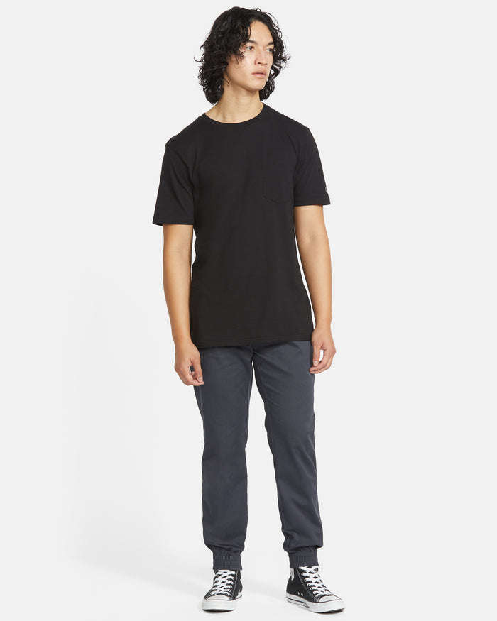 A person with medium-length curly hair stands sideways, wearing a black t-shirt, Volcom Men's Frickin Slim Joggers, and black-and-white sneakers against a plain white background.