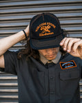 A man in front of a metal shutter wears the Volcom Men's Fundamentals Work Hat and a dark cotton twill shirt, both featuring orange embroidered logos. He holds the hat's brim and looks down.