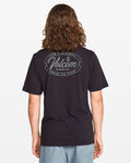 A person with long brown hair, facing away, wears the Volcom Men's Garaged Short Sleeve Tee in washed black heather with a circular “The Original Volcom Stone Co. True To This” back design, paired with light gray shorts.